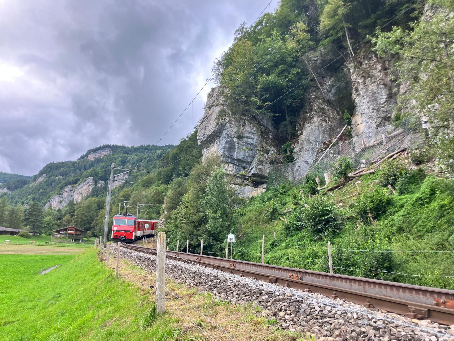 A red train approaching along tracks beneath a forested rock face